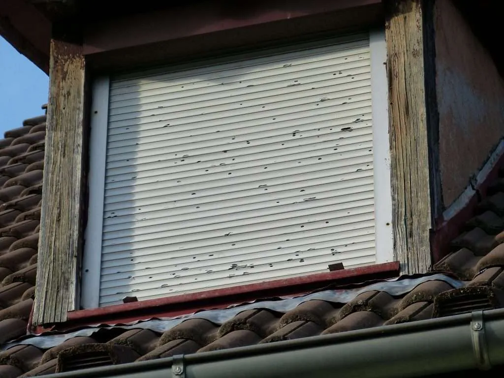 Metal storm shutters show signs of horizontally blown hail and potential roof damage.